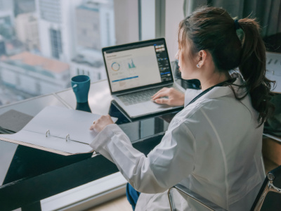 Female doctor in white coat sitting at a desk and looking at laptop and binder with graphs and charts